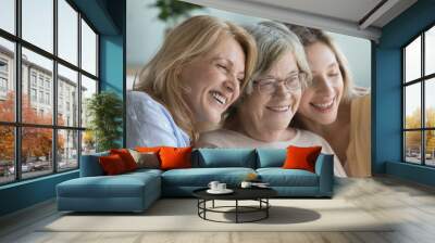 Cheerful ladies of three female generations meeting at home, hugging, looking away with toothy smiles. United family portrait of older grandmother, mother and young adult daughter woman. Close up Wall mural