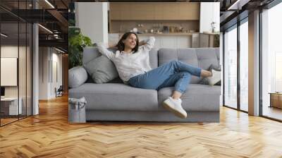 Cheerful millennial woman relax on sofa in studio apartment, leaned on soft comfortable couch with hands behind head, smile enjoy carefree leisure at smart home with climate control for comfort life Wall mural