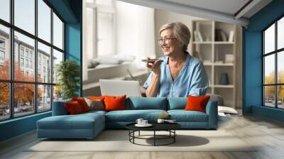 Cheerful older pensioner woman using gadgets for wireless online communication at home, smiling, laughing at table with laptop, holding smartphone at face, recording message Wall mural