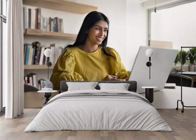 Cheerful successful young Indian project manager woman enjoying online communication at computer, typing on laptop in co-working space, smiling, working, using job application, browsing Internet Wall mural