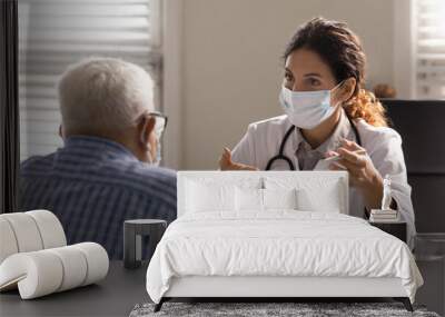 Close up female therapist wearing face mask consulting mature patient at meeting in hospital office, doctor talking, explaining, discussing medical checkup result or symptoms with older man Wall mural