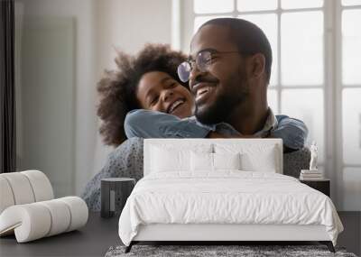 Close up happy African American father wearing glasses piggy backing little daughter, engaged in funny activity, standing at home, cute girl embracing overjoyed dad, family having fun together Wall mural