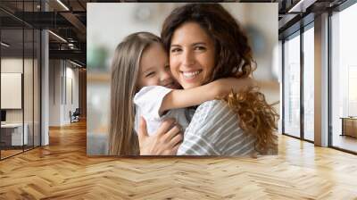 Close up headshot portrait picture of smiling mother hugging daughter sitting on couch at home. Happy young parent with little child embracing looking at camera posing for photo in living room. Wall mural