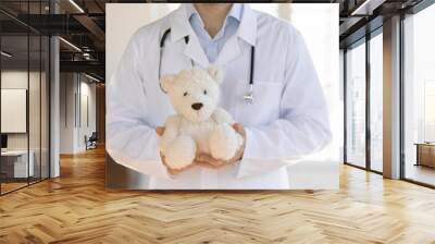 Close up male doctor pediatrician holding little patient fluffy toy teddy bear in hands, therapist gp wearing white coat uniform with stethoscope standing in clinic, children healthcare concept Wall mural