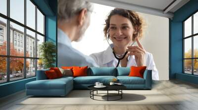 Close up of smiling young Caucasian female doctor use stethoscope examine mature patient heart rate in clinic. Happy caring woman nurse hold phonendoscope listen to elderly man heartbeat in hospital. Wall mural