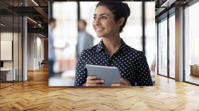 Close up smiling dreamy Indian businesswoman holding computer tablet, looking to aside, intern thinking about new job opportunities, startup ideas, dreaming about good future, business success Wall mural