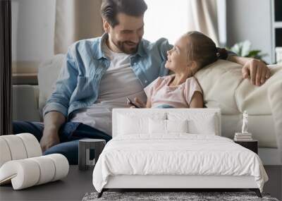 Close up smiling father and little daughter using phone together, sitting on cozy couch at home, happy young dad and adorable child girl enjoying leisure time, weekend in living room Wall mural