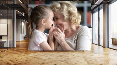 Close up smiling grandmother and little granddaughter talking, laughing, pretty preschool girl sharing secrets with happy grandma, whispering, having fun, sitting on cozy couch at home Wall mural