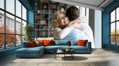 Close up smiling mature woman wearing glasses hugging adult son, standing in living room at home, family enjoying tender moment, beautiful happy middle aged woman embracing young man Wall mural