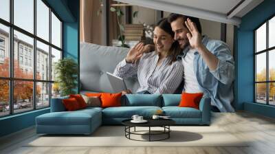 Close up smiling young couple using tablet, sitting on couch at home together, happy woman and man waving hands, greeting, using webcam, chatting online, making video call to relatives or friends Wall mural