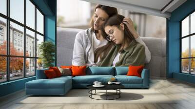 Compassionate young mother comforting depressed sad teen girl, hugging daughter with love, care, speaking, giving sympathy, advice, support, holding kids hand, sitting on home couch Wall mural