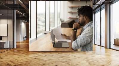 Concentrated young african man read printed document before laptop screen compare data think on text edit check errors. Thoughtful black male analyse financial report plan changes in contract project Wall mural