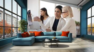 Concentrated young indian employee showing marketing research data to focused blonde supervisor in glasses. Two mixed race colleagues developing economic growth strategy, analyzing graphs on computer. Wall mural