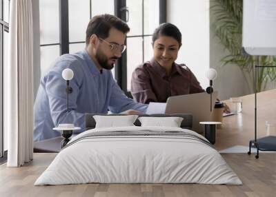 Confident businessman mentor teaching smiling Indian intern, reading documents, diverse colleagues sitting at desk with laptop, working on project together, manager consulting client at meeting Wall mural