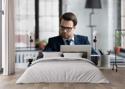 Confident businessman wearing suit and glasses using laptop, writing notes or financial report, sitting at desk in office, focused young entrepreneur working with documents or marketing plan Wall mural