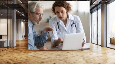 Confident young 30s female general practitioner physician showing medical insurance program presentation or health lab test results on computer to focused middle aged older patient at clinic meeting. Wall mural