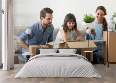 Cute kid daughter helping mom dad unpack boxes in new home on moving day concept, happy family of three child girl and parents packing belongings relocating sitting on sofa in living room together Wall mural