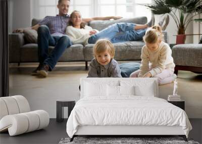 Cute kids playing while parents relaxing sofa at home together, smiling active boy entertaining with toy car near his sister on floor, happy family spending time together in living room on weekend Wall mural