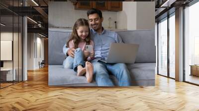 Cute preschool daughter holding smartphone sit on sofa with young father use laptop, family using diverse devices spend leisure on internet. Young generation and gadgets overuse, apps usage concept Wall mural