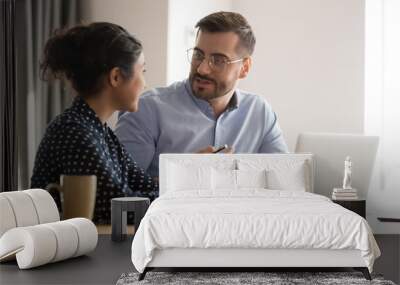 Different ethnicity millennial co-workers indian woman and caucasian man sit at desk discuss new project or task, share information brainstorm creative innovative ideas, teamwork and thinking concept Wall mural