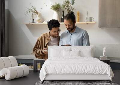 Distracted from cooking happy young African American family couple using cellphones, checking interesting food recipe, communicating online, inviting friends for party, preparing meal in kitchen. Wall mural