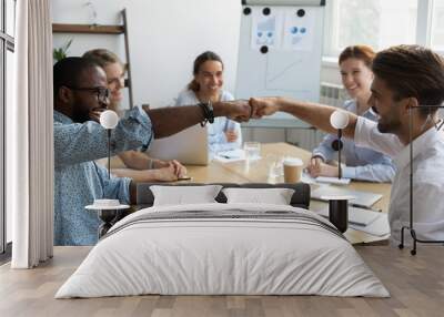 Diverse company staff girls guys sitting at desk in boardroom feel happy and satisfied celebrating success at work. Diverse colleagues fist bumping greeting each other express friendship and respect Wall mural