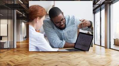 Diverse millennial colleagues working together analyzing diagram looking at computer screen. Black mentor helps female apprentice understand corporate program explaining interface showing on monitor Wall mural