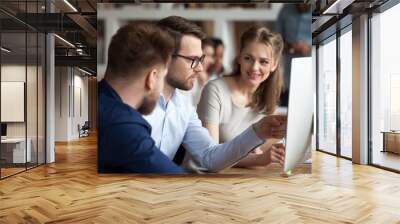 Diverse millennial employees coworking discussing business project or idea in shared office, smiling colleagues work together at pc, male worker explain to coworkers pointing at computer screen Wall mural