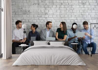 Diverse millennial people with different gadgets talking and laughing, sitting on chairs in row, using laptops, tablets, mobile phones, sharing and discussing content on screen, learning new apps Wall mural