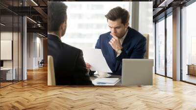 Doubting businessman reading contract document with suspicion when sitting at desk in front of business partner. Entrepreneur unsure in terms of agreement, feeling skeptical because of financial plan Wall mural