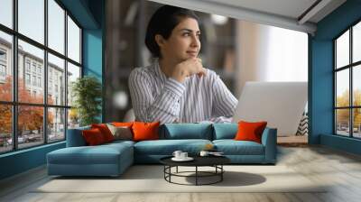 Dreamy young Indian woman sit at desk at home office work on laptop thinking or pondering. Pensive millennial mixed race female distracted from computer job look in distance visualizing planning. Wall mural