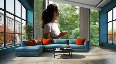 Dreamy young woman in eyeglasses looking out of opened window, breathing fresh air, holding smartphone in hands. Happy millennial lady daydreaming, visualizing future, feeling inspired indoors. Wall mural