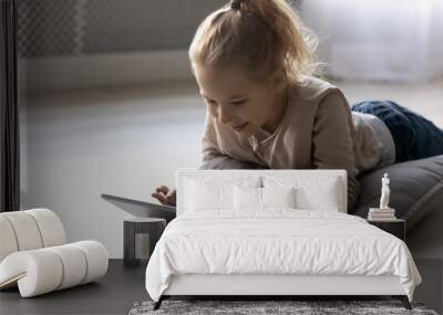 Emotional positive happy little kid girl involved in playing online games on digital computer tablet, lying on comfortable soft pillow on warm wooden floor, communicating online in social network. Wall mural