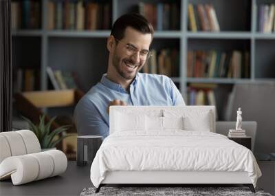 Excited man wearing glasses celebrating success, reading good news in email, happy overjoyed businessman looking at laptop screen, showing yes gesture and laughing, sitting at work desk Wall mural