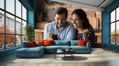 Excited millennial Caucasian man and woman feel euphoric win online lottery on smartphone. Happy overjoyed young couple look at cellphone screen triumph with good news or deal message. Wall mural