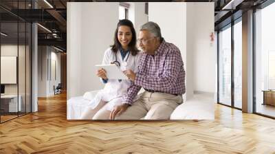 Excited old Indian patient man and happy young doctor woman using digital tablet together, looking at display, talking, discussing medical test results, modern electronic diagnosing Wall mural