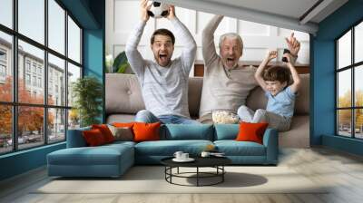 Excited three generations of men sport fans relax in living room celebrate team victory together, overjoyed little boy with dad and grandfather have fun watching football match at home together Wall mural