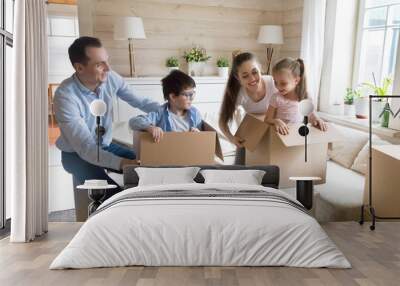 Excited young family have fun together while unpacking moving in, playful cute kids hide in boxes when opening packages with belongings, happy parents play game with children relocating to new house Wall mural