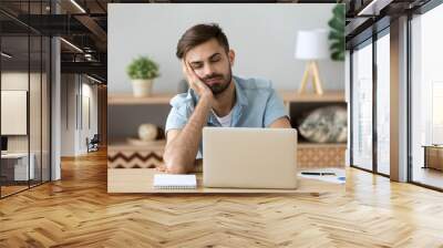 Exhausted millennial male hold head with hand sitting at office table falling asleep, tired man fall asleep working at laptop at home workplace, sleepy guy feel fatigue taking nap near computer Wall mural