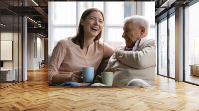 Family members of two generations grown daughter and mature father having fun enjoy tea talk on cozy couch. Attentive young lady caregiver social worker visit support care for glad elderly man patient Wall mural