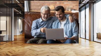 Family time with grown son. Elder and younger men relatives sit on sofa at home hug look on laptop screen discuss online news. Retired dad interested in pc talk ask adult child question about app work Wall mural