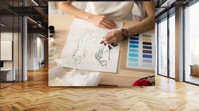 Female hands holding brush painting embroidery pattern scheme on fashion sketch, designer drawing new mens womens wear winter collection sampler at desk with color swatches palette, close up view Wall mural