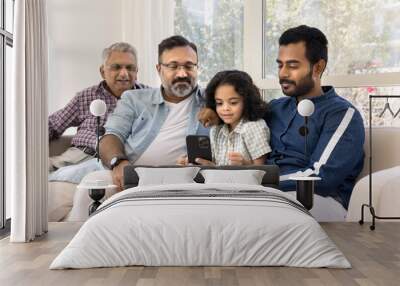 Focused Indian relatives of four different family generations using online application, media service for entertainment on cellphone at home, sitting on sofa together Wall mural