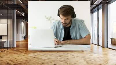 Focused man wearing headphones writing notes studying with laptop, serious student or office worker listening to audio business course at work, e-learning and online professional education concept Wall mural