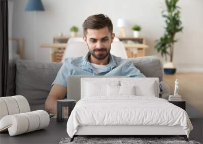 Focused millennial man sit on couch in apartment using laptop browsing internet, young male relax on sofa busy with computer surfing web or reading news, man work from home outsourcing or networking Wall mural