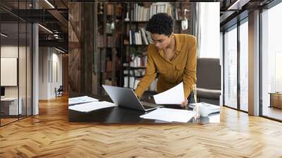 Focused serious young Black businesswoman holding, checking documents, working at laptop computer. African worker, professional using computer in home office, doing paperwork job, standing at table Wall mural