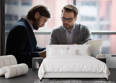 Focused young businessman signing agreement with skilled lawyer in eyeglasses. Concentrated financial advisor showing place for signature on paper contract document to male client at meeting in office Wall mural