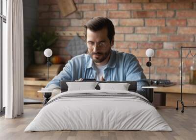 Focused young Caucasian man sit at desk at home look at smartphone screen having online consultation on device. Serious male use cellphone text or message on internet. Technology concept. Wall mural