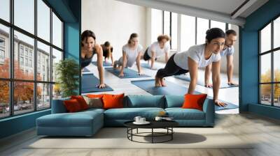 Focused young multiracial people practicing plank position on straight arms with professional indian ethnicity trainer at group class, strengthening back or abdominal core muscles together indoors. Wall mural