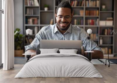 Front view head shot young smiling african american businessman in eyewear looking at laptop screen, reading pleasant messages in social networks, working on computer sitting at table in office home. Wall mural